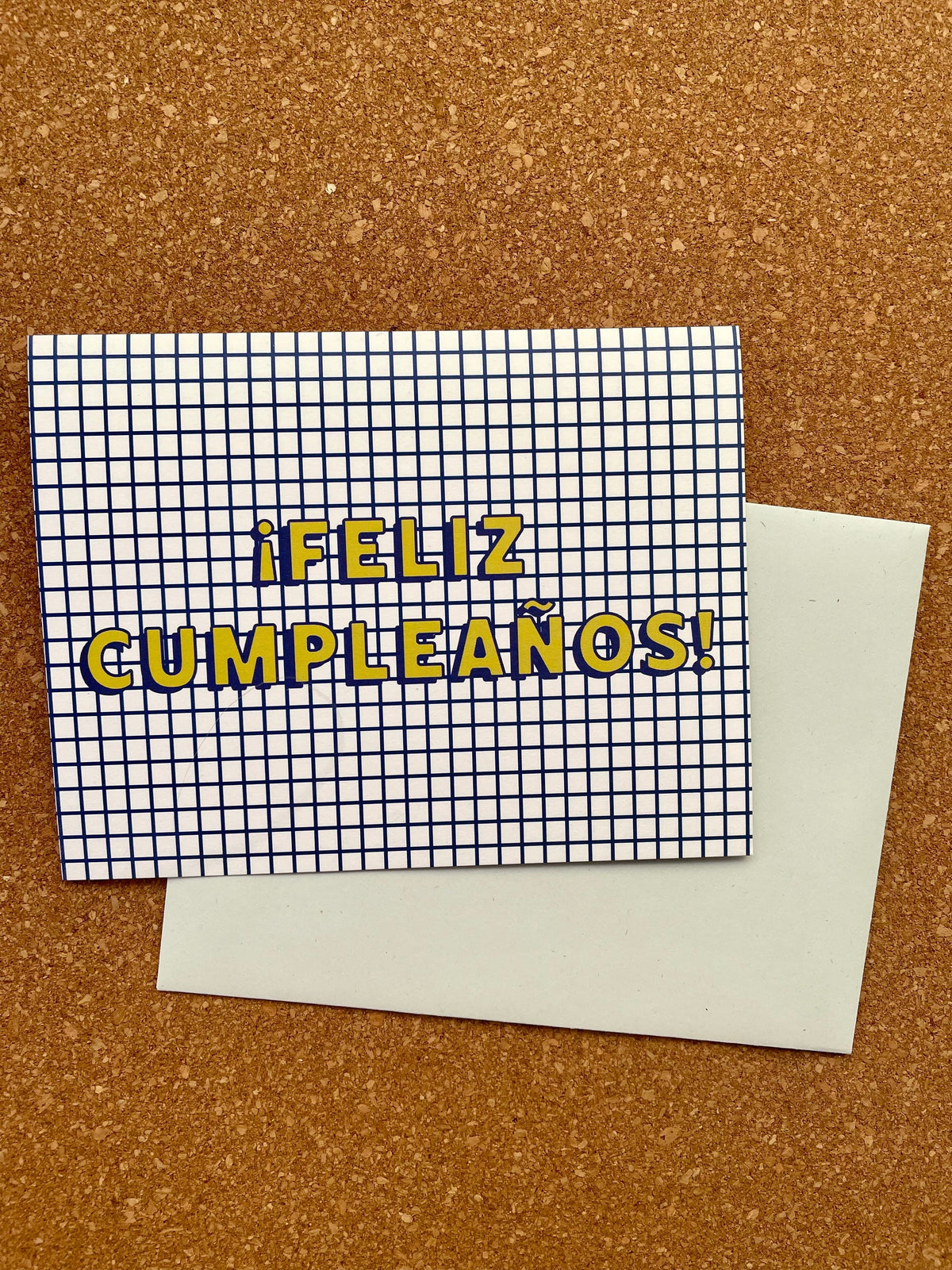 Greeting card with 'Feliz Cumpleaños!' printed in yellow on a grid background, paired with a pale blue envelope, presented on a cork backing.