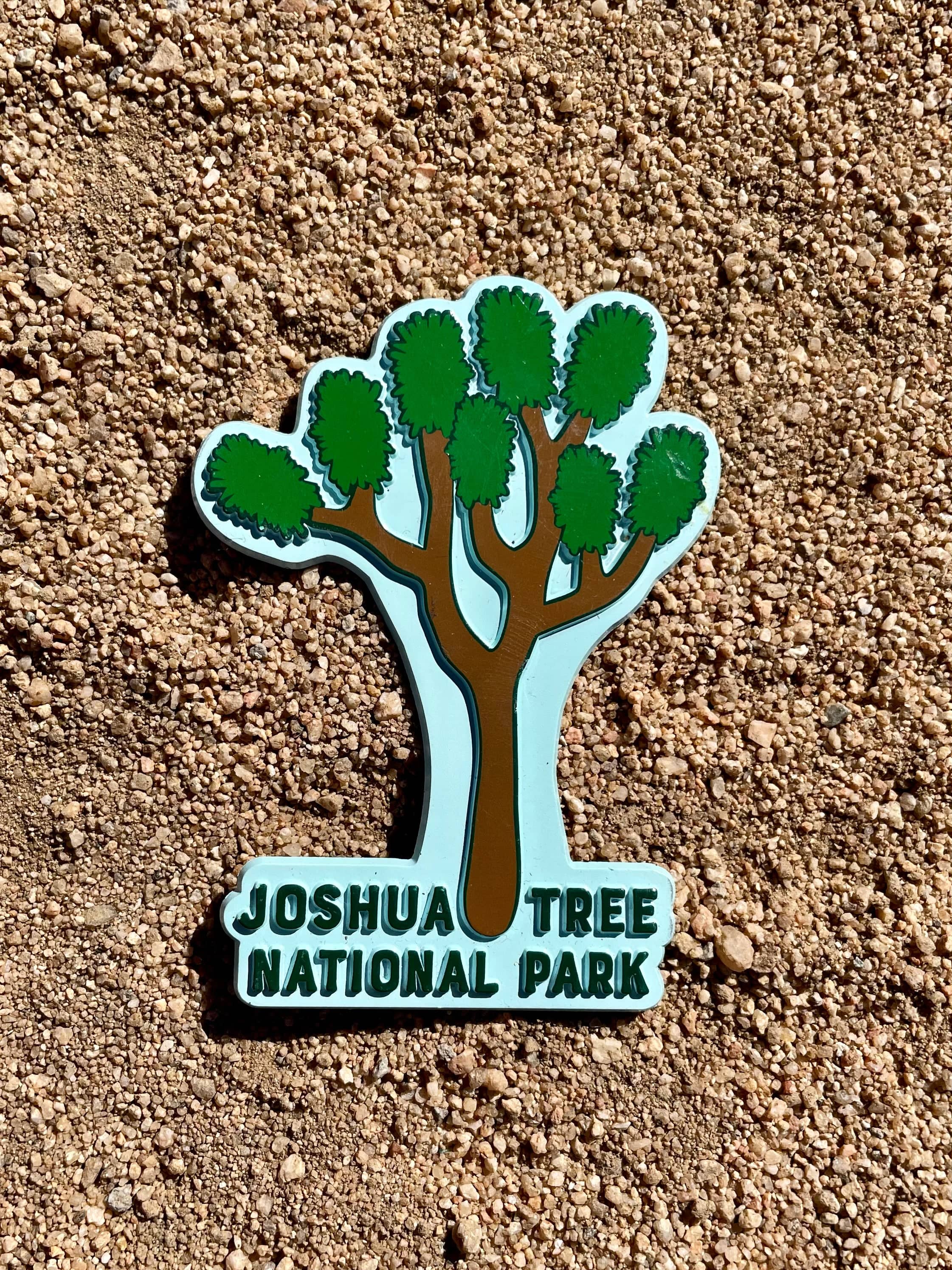 A rubber magnet with the shape of Joshua Tree National Park, featuring a stylized tree and text in green on a light blue background.