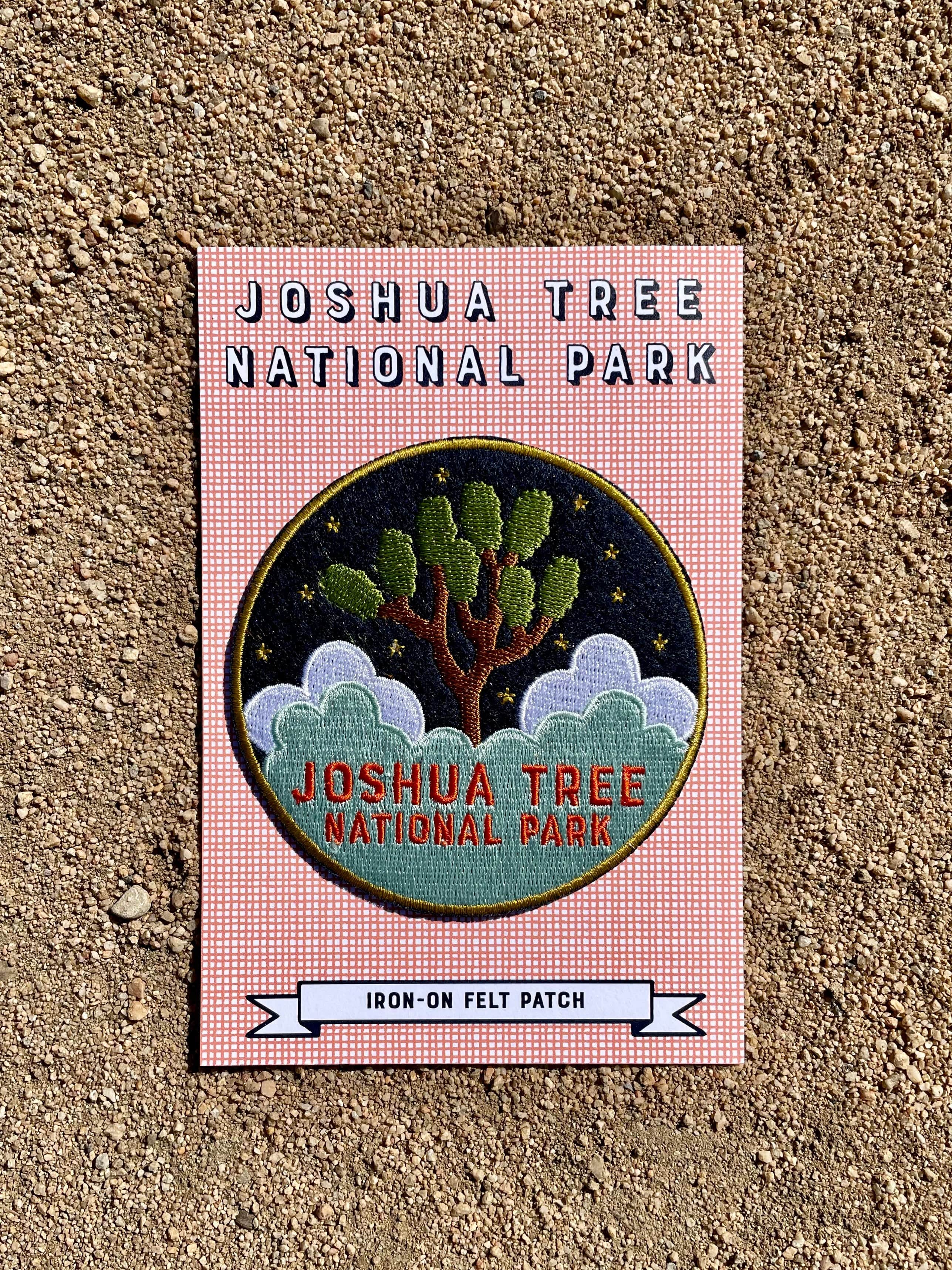 A round navy blue felt patch with embroidered white text and imagery related to Joshua Tree National Park, placed on a cardboard backing with printed text.
