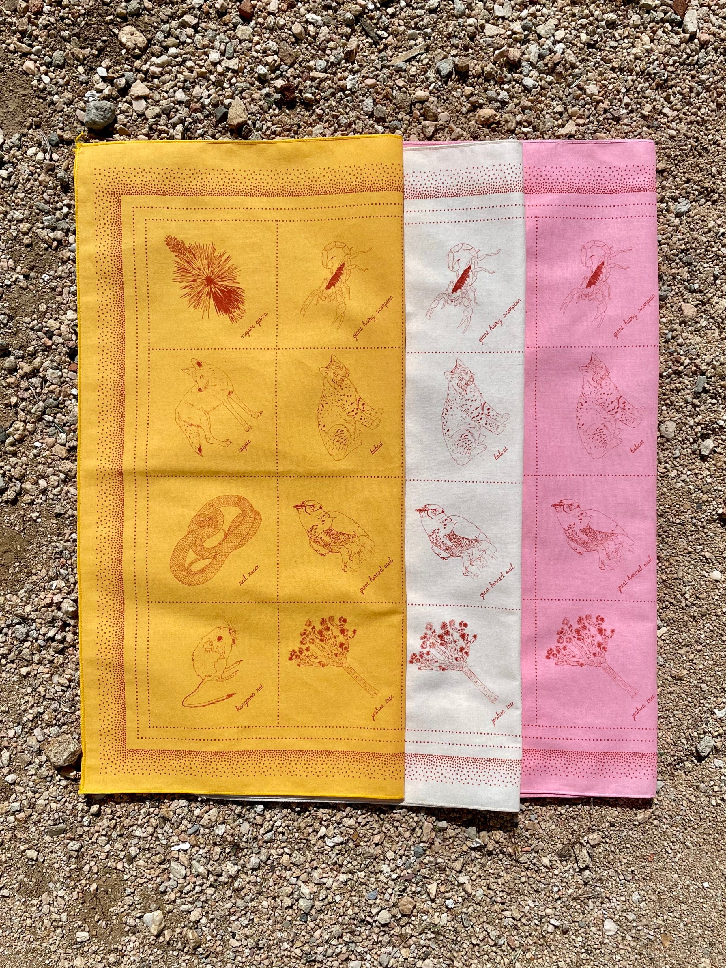 Two bandanas laid out on a surface, one yellow and one pink, both with brown floral and animal print designs.