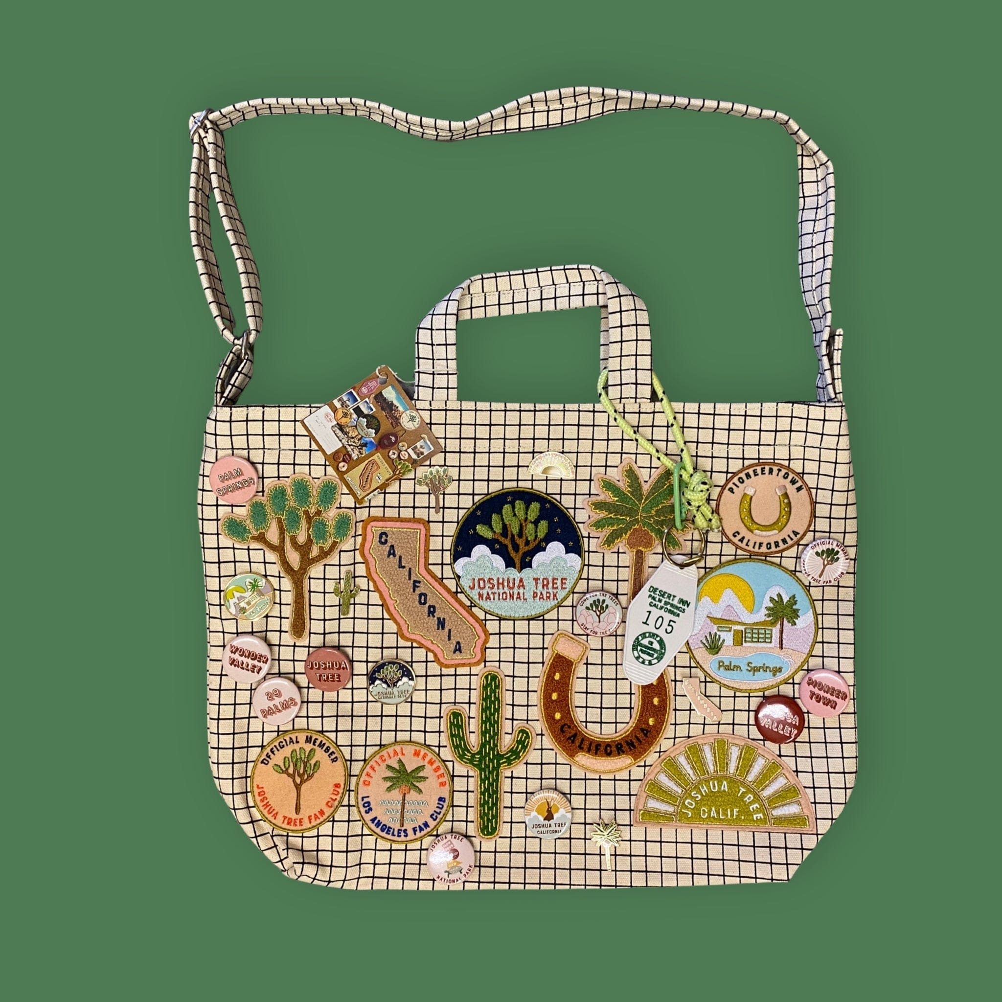 A beige and white checkered canvas shoulder bag decorated with various colorful embroidered patches, charms, and appliqués, set against a solid green background.