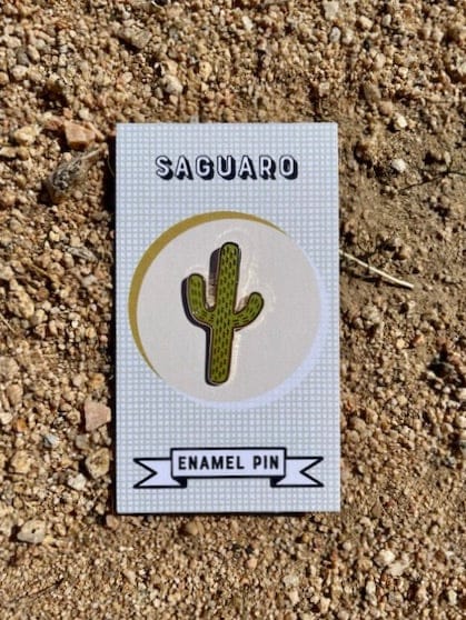 An enamel pin shaped like a saguaro. It is green with gold details.