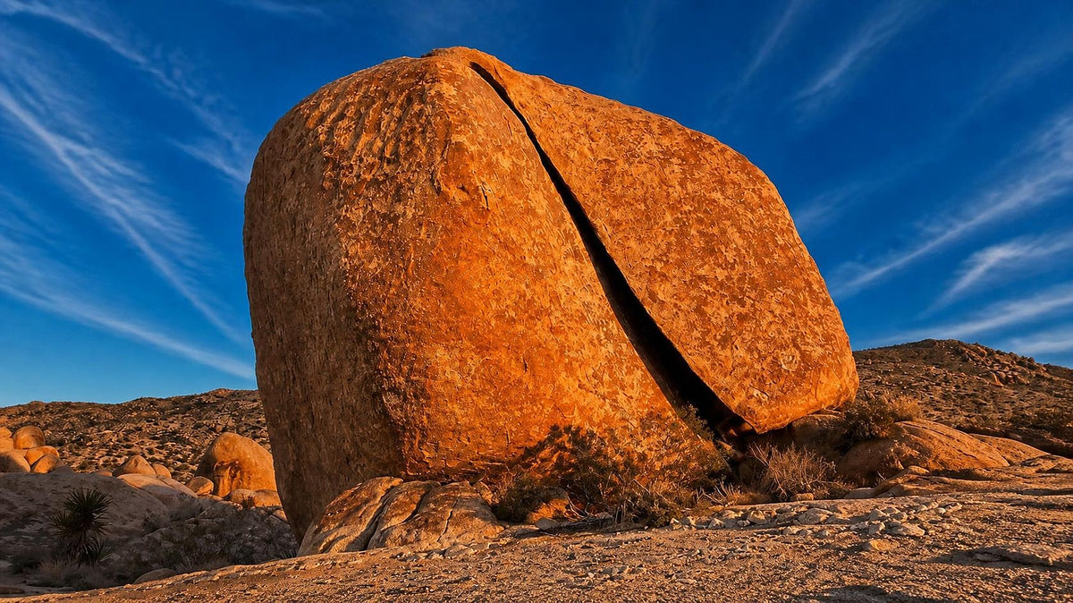Split Rock in Joshua Tree National Park, a huge rounded monzogranite boulder cleaved by a deep vertical fracture, glowing orange at sunset under a deep blue sky