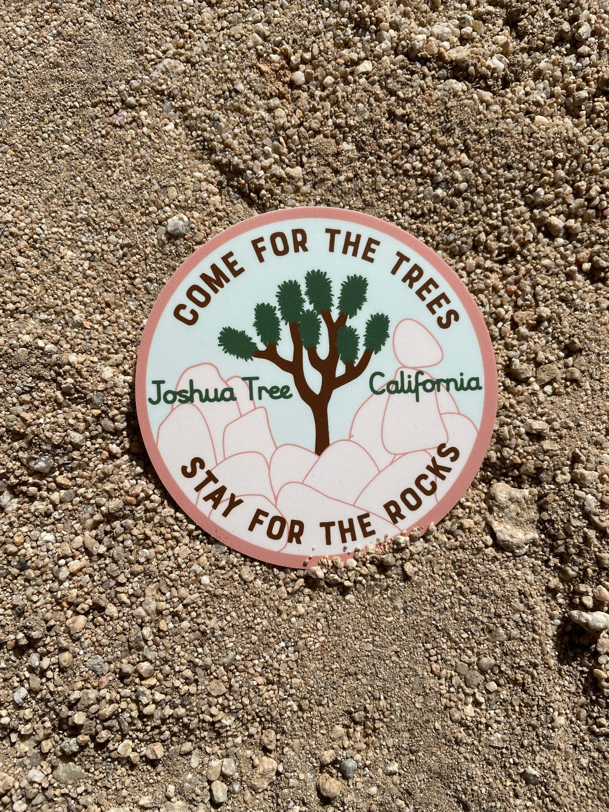 A sticker with a tree and the text 'JOSHUA TREE, CALIFORNIA. STAY FOR THE ROCKS.' on it, placed on a ground surface.