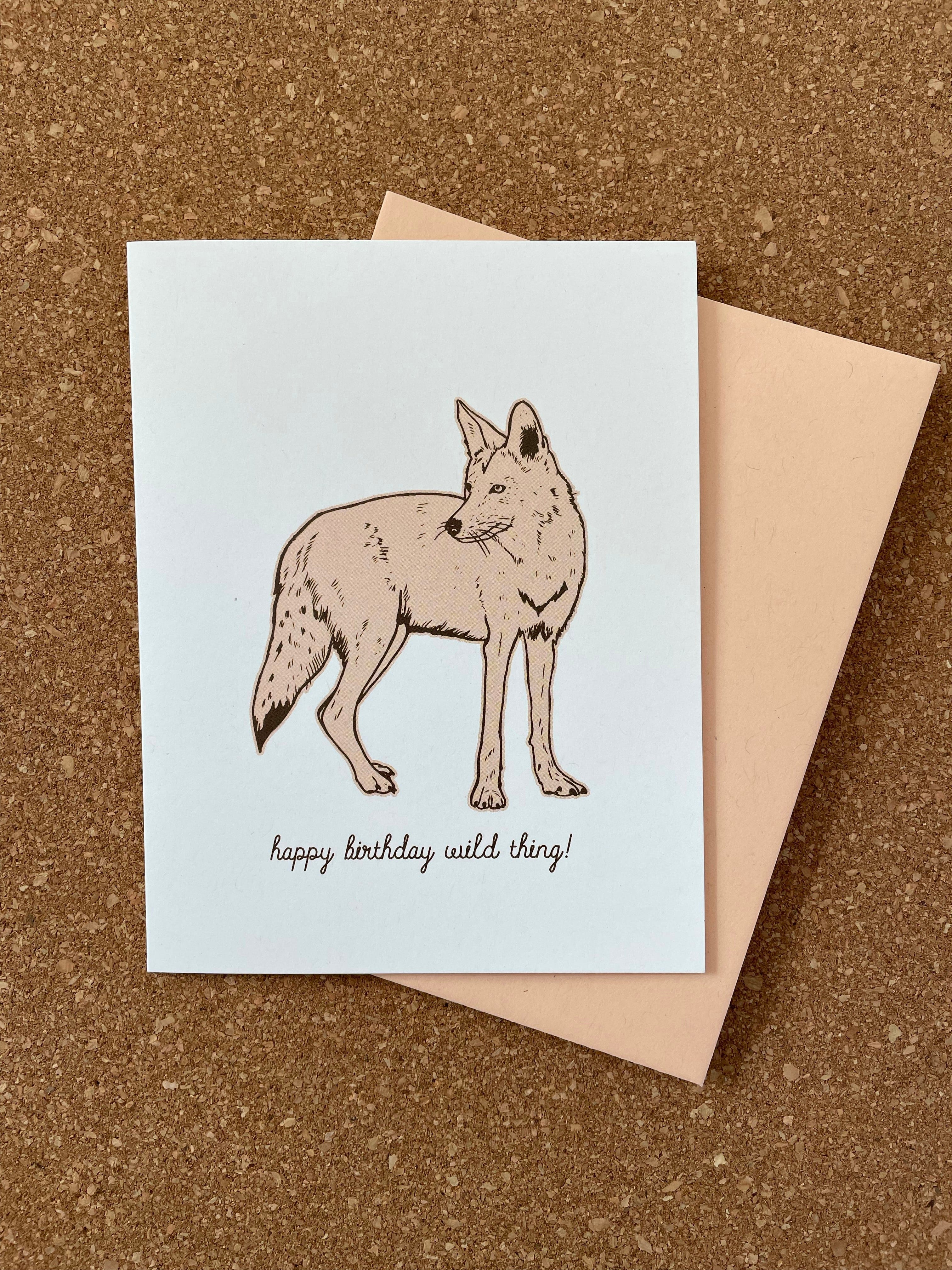 Greeting card with an illustration of a coyote and the text 'happy birthday wild thing!' next to an orange kraft-tone envelope, all placed on a cork background.