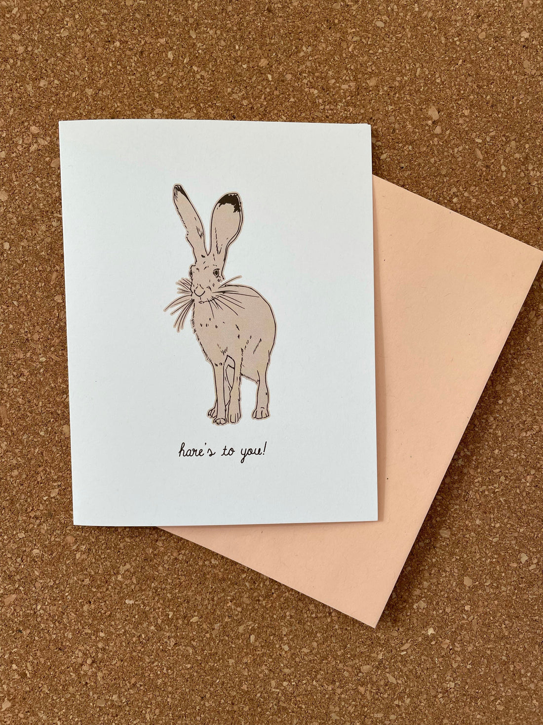 A greeting card with a line drawing illustration of a hare, accompanied by an orange kraft-tone envelope, placed on a cork background.