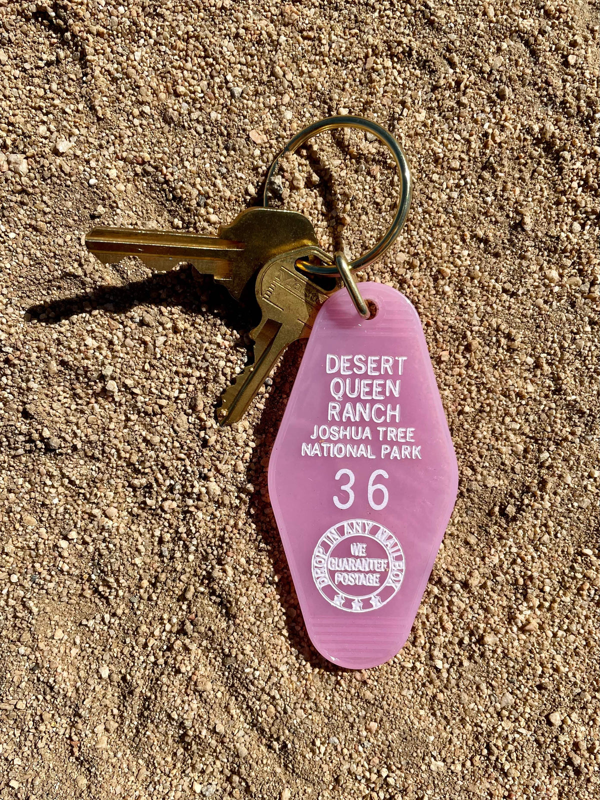 Authentic motel keychain with translucent pink key tag and brass keyring, featuring the text 'Desert Queen Ranch Joshua Tree National Park' and the number '36', placed on a sandy surface.