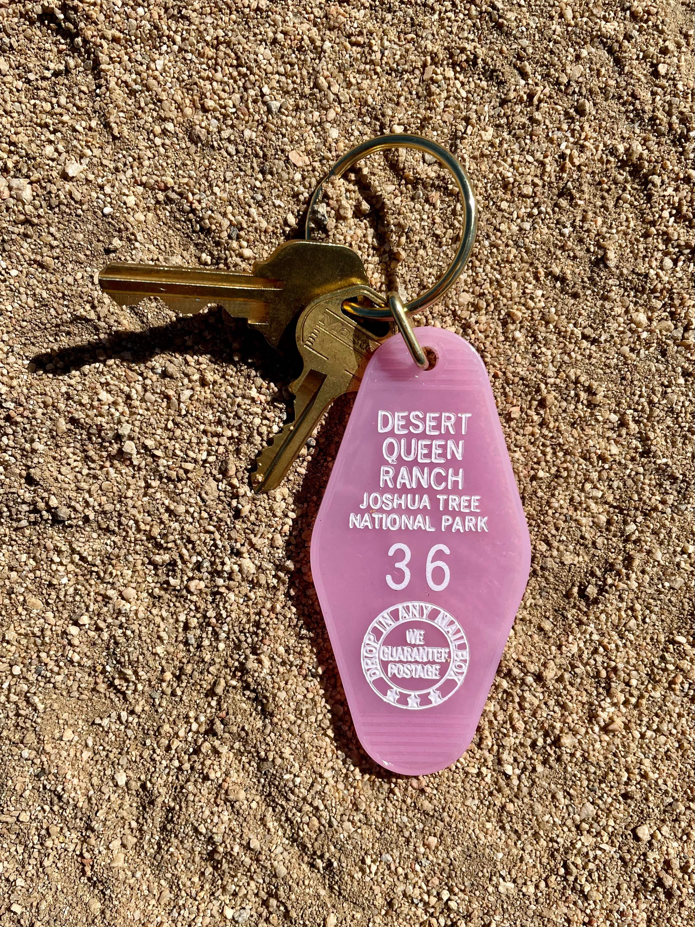 Authentic motel keychain with translucent pink key tag and brass keyring, featuring the text 'Desert Queen Ranch Joshua Tree National Park' and the number '36', placed on a sandy surface.