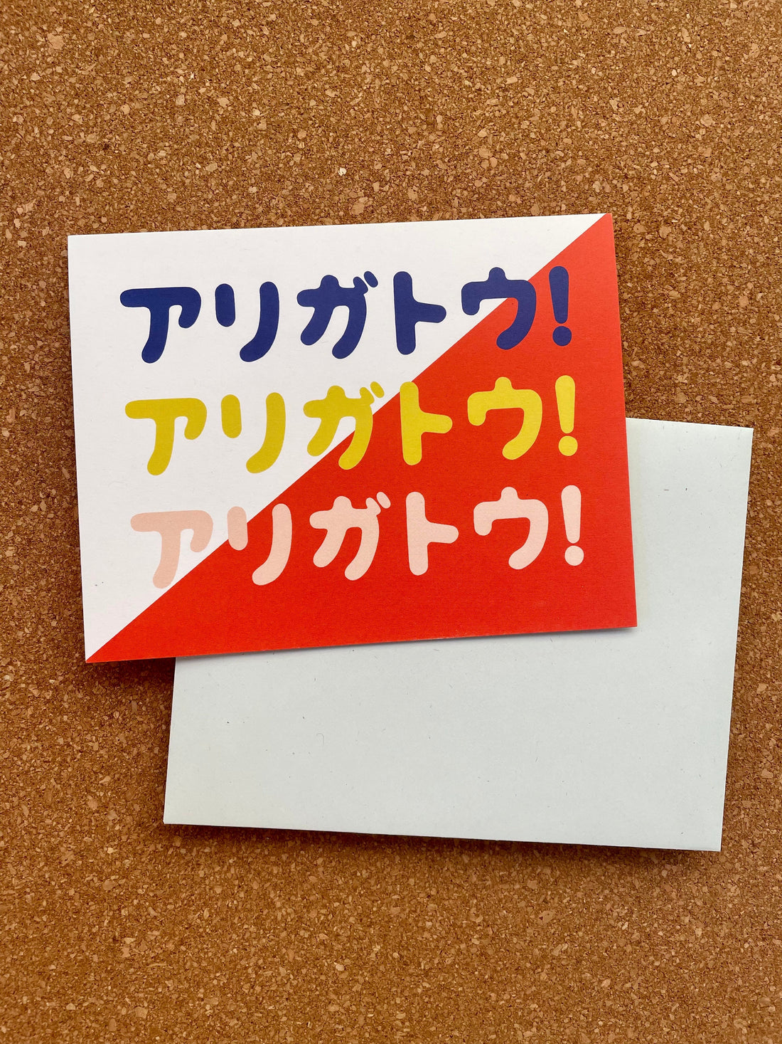 A greeting card with a red and blue design and the word 'Arigato' printed on it in katakana accompanied by a pale blue envelope.