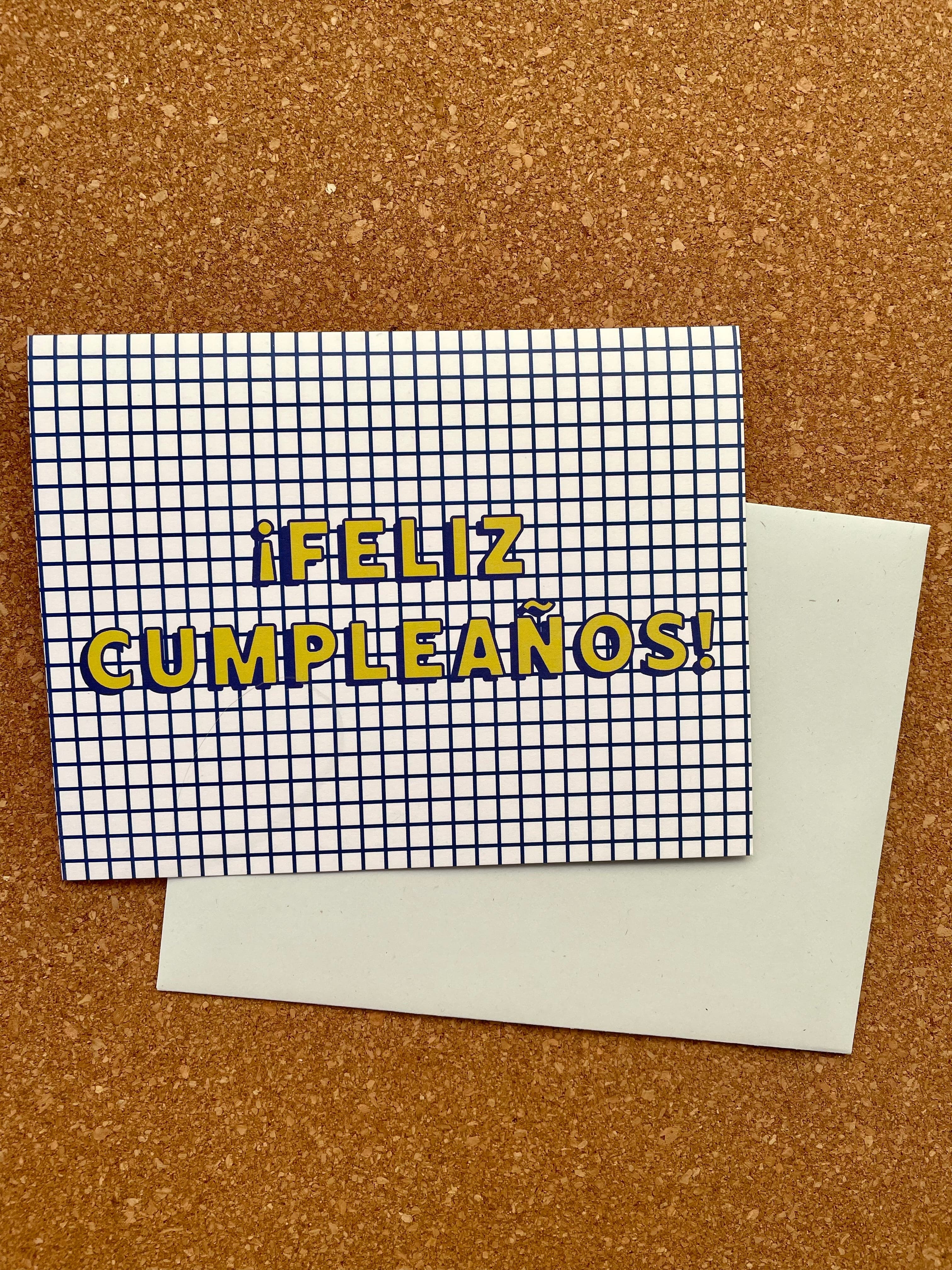 Greeting card with 'Feliz Cumpleaños!' printed in yellow on a grid background, paired with a pale blue envelope, presented on a cork backing.