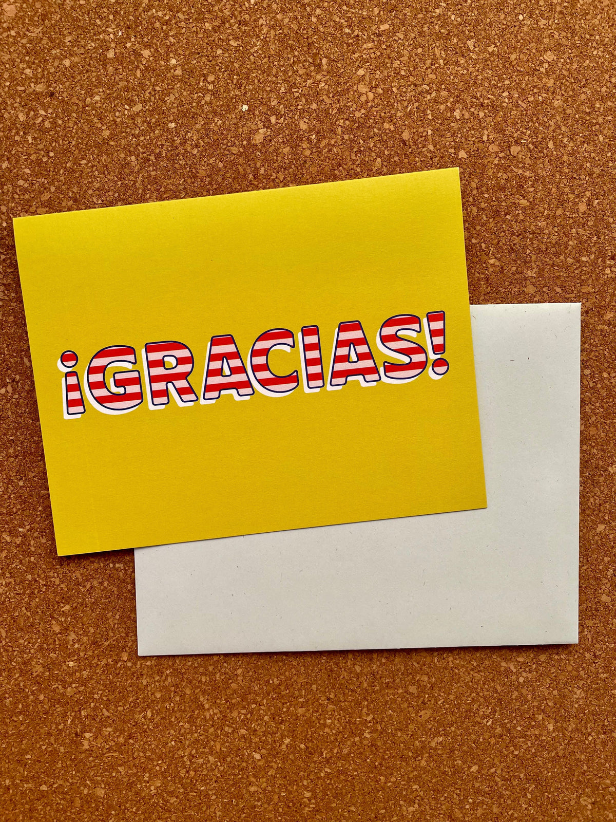 A yellow greeting card with the text 'GRACIAS!' in red, placed on a cork board background with a pale blue envelope beside it.