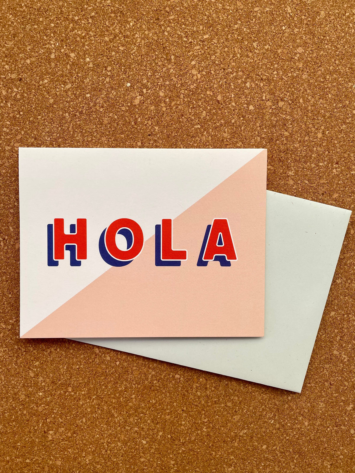 A pink and white greeting card with the text 'HOLA' in red, placed on a cork board, with a pale blue envelope beside it.