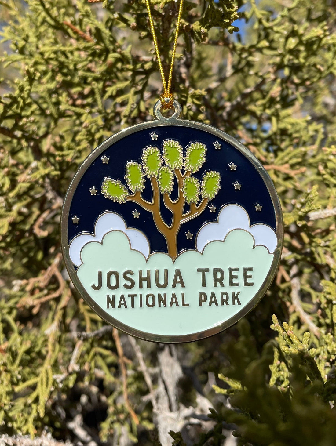 A round enamel ornament featuring the Joshua Tree National Park emblem with gold metal and a gold ribbon, displayed against a Christmas tree backdrop.