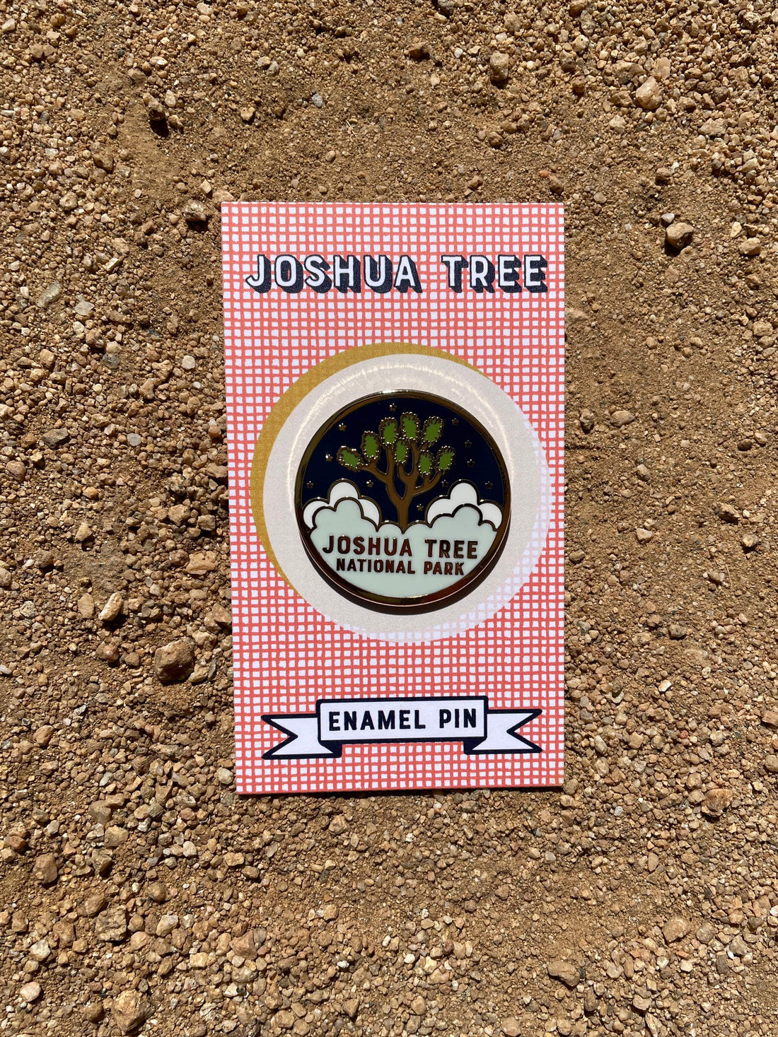 A round enamel pin with a gold metal and hard enamel design featuring the text 'JOSHUA TREE NATIONAL PARK' and an illustration of a tree, placed on a dirt surface.