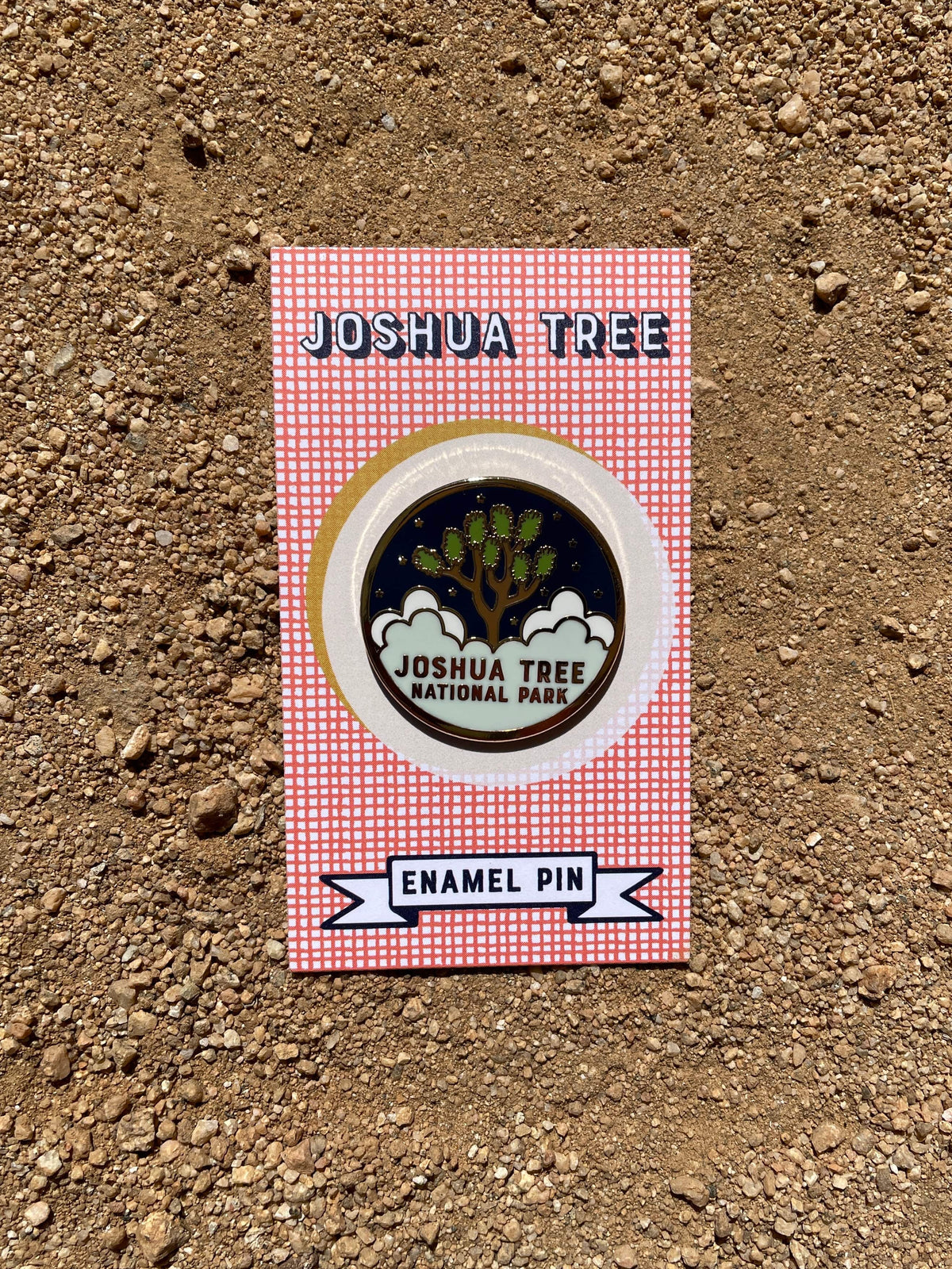 A round enamel pin with a gold metal and hard enamel design featuring the text 'JOSHUA TREE NATIONAL PARK' and an illustration of a tree, placed on a dirt surface.