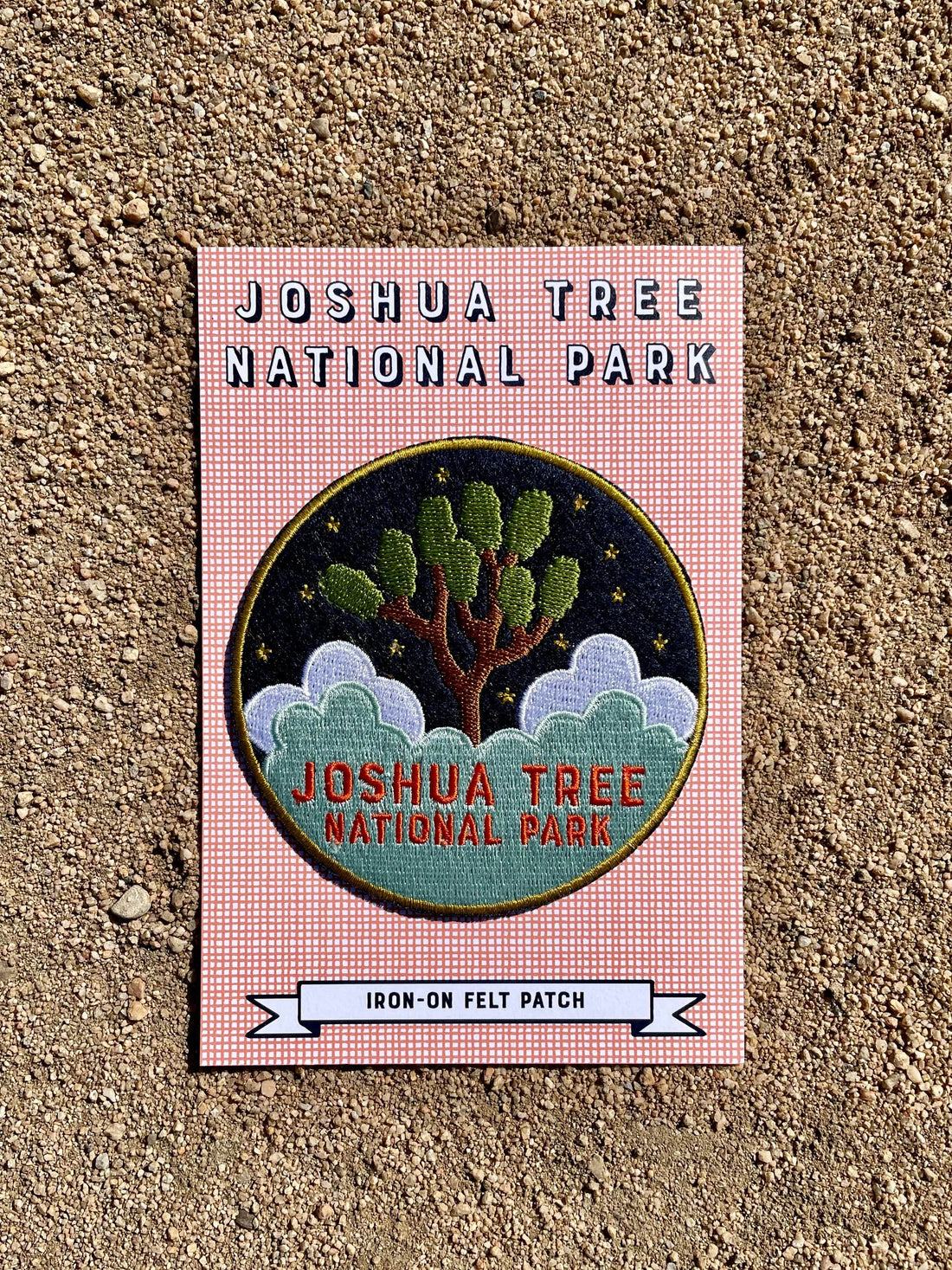 A round navy blue felt patch with embroidered white text and imagery related to Joshua Tree National Park, placed on a cardboard backing with printed text.