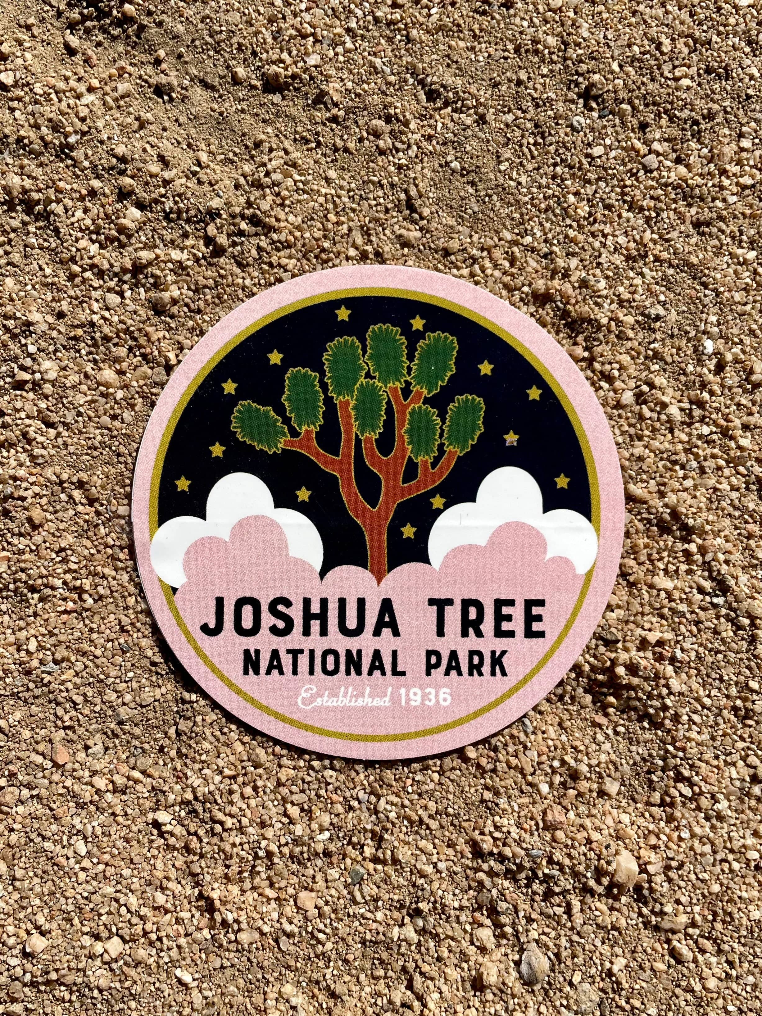 A round sticker with an illustration of Joshua Tree National Park, predominantly featuring a tree with green leaves, against a black background with pink at the bottom.