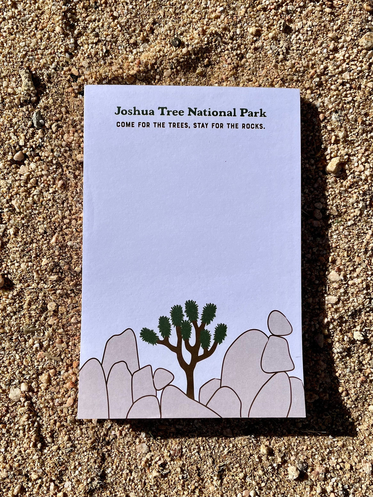 A 50-page notepad with a white cover, featuring a small illustration of a Joshua tree and rocks, lying on a sandy surface.