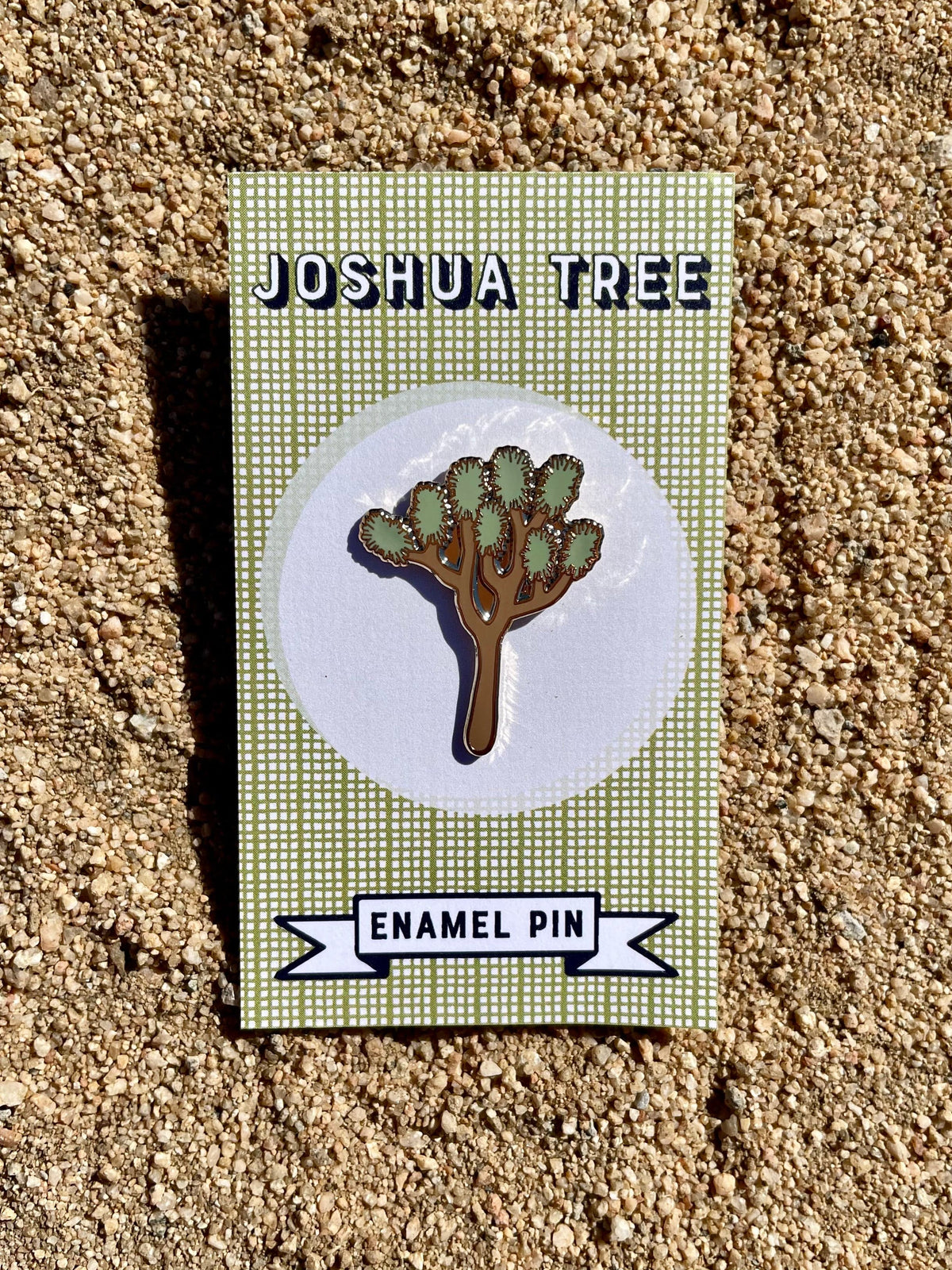 An enamel pin shaped like a Joshua Tree displayed on a backing card with the text 'JOSHUA TREE ENAMEL PIN'. The pin is gold-colored with a white rubber pinback.
