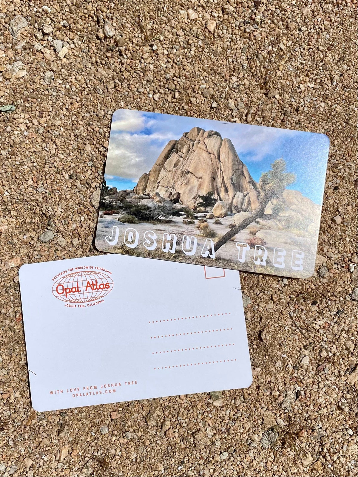 A 4x6 inch matte postcard with rounded corners displaying an image of Old Woman Rock in Joshua Tree National Park, lying on a sandy surface.