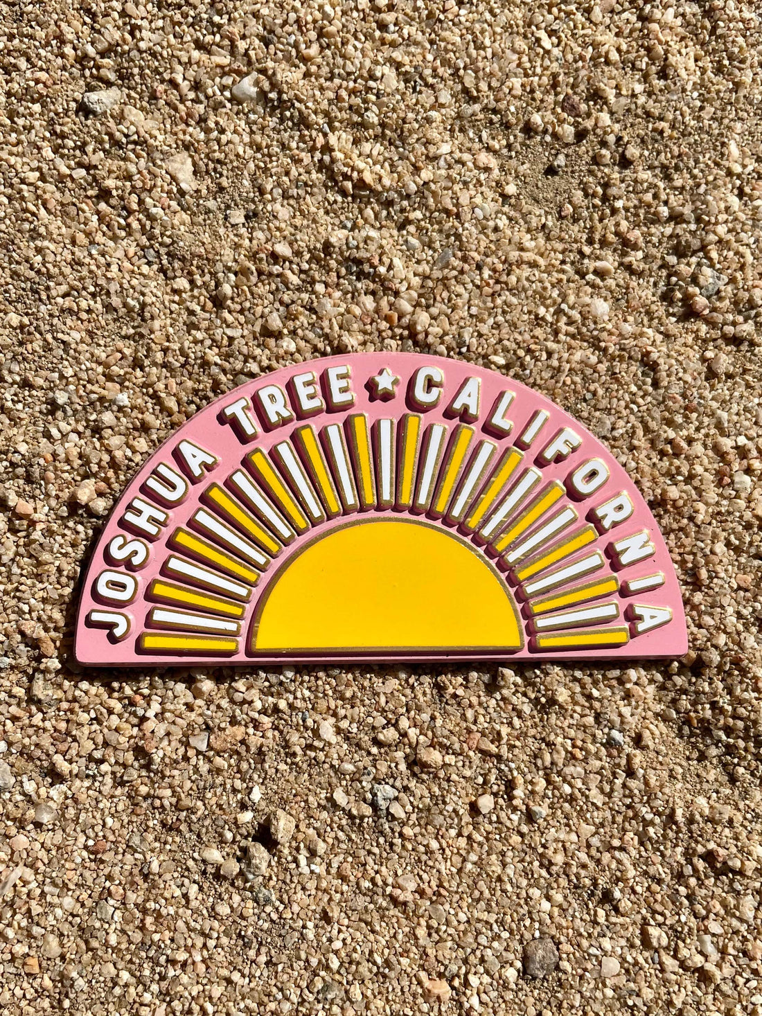 A rubber magnet with a design resembling a sun and the text 'JOSHUA TREE CALIFORNIA' on a pink background, placed on a sandy surface.