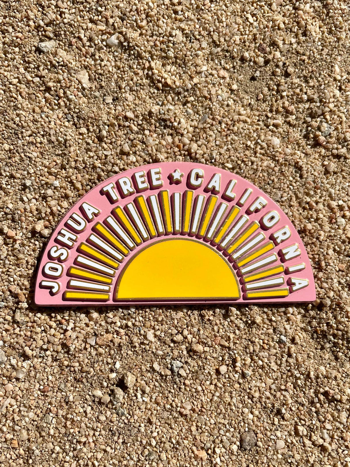 A rubber magnet with a design resembling a sun and the text 'JOSHUA TREE CALIFORNIA' on a pink background, placed on a sandy surface.