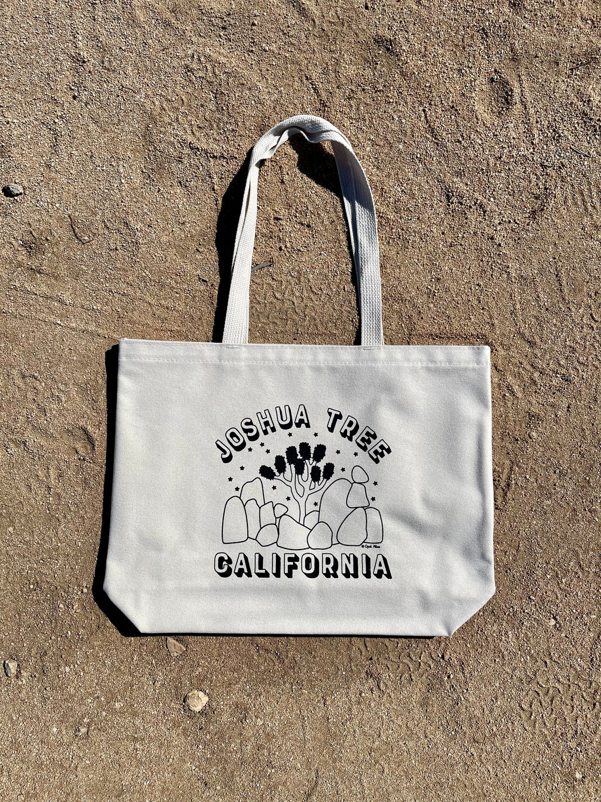 A beige cotton tote bag with 'Joshua Tree California' printed in black text on the front, laid out on a sandy surface. 