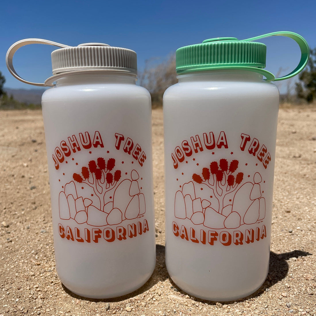 Two white Nalgene water bottles with red 'Joshua Tree California' text and a cactus design, one with a gray cap and one with a green cap, on a sandy surface with a clear sky in the background.