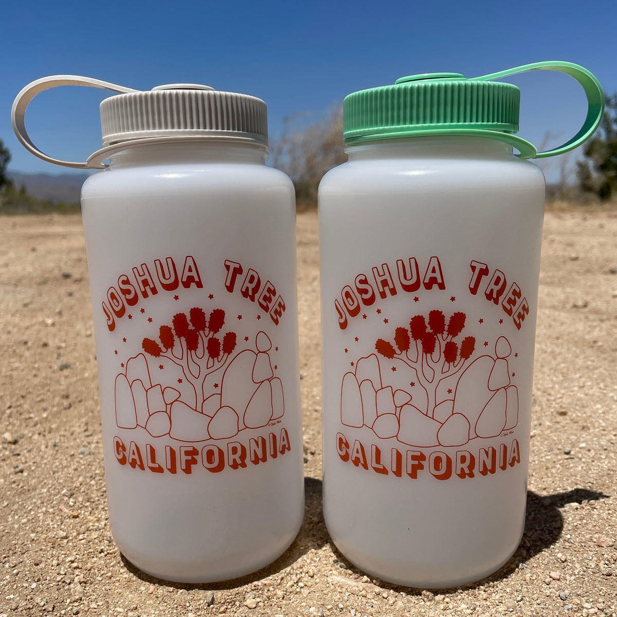 Two white Nalgene water bottles with red 'Joshua Tree California' text and a cactus design, one with a gray cap and one with a green cap, on a sandy surface with a clear sky in the background.