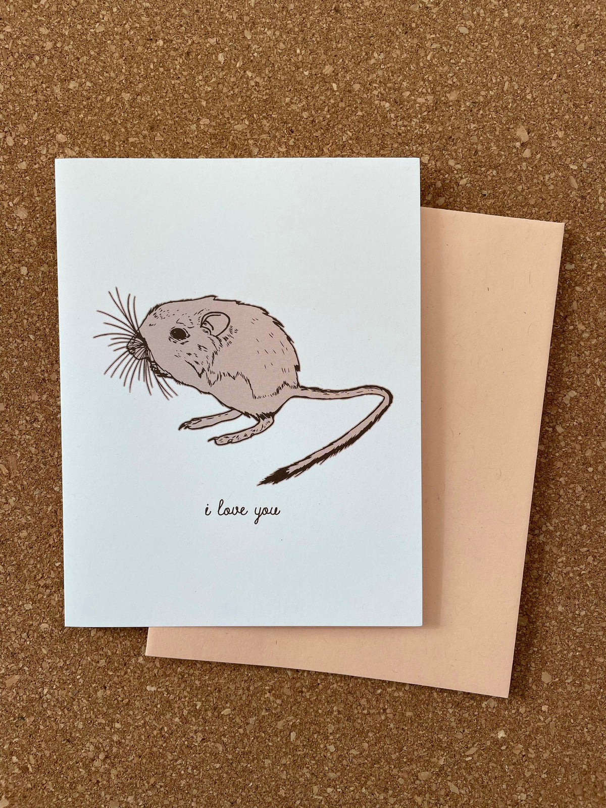 Greeting card with an illustration of a kangaroo rat and the phrase 'i love you' printed on it, accompanied by an orange kraft-tone envelope, all placed on a cork background.
