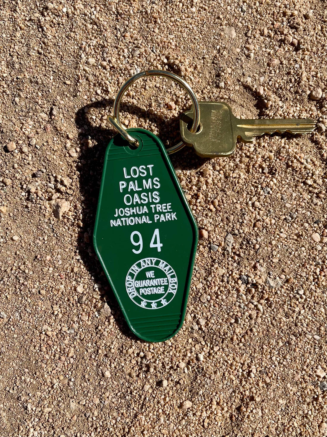 A green motel-style keychain with the inscription 'LOST PALMS OASIS JOSHUA TREE NATIONAL PARK' and the number '94', stamped on the tag, lying on a sandy surface.