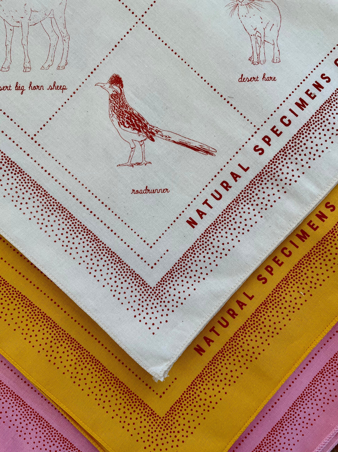 Detail of natural specimens bandana, showing the roadrunner in red ink on natural colored bandana with gold and pink versions underneath.