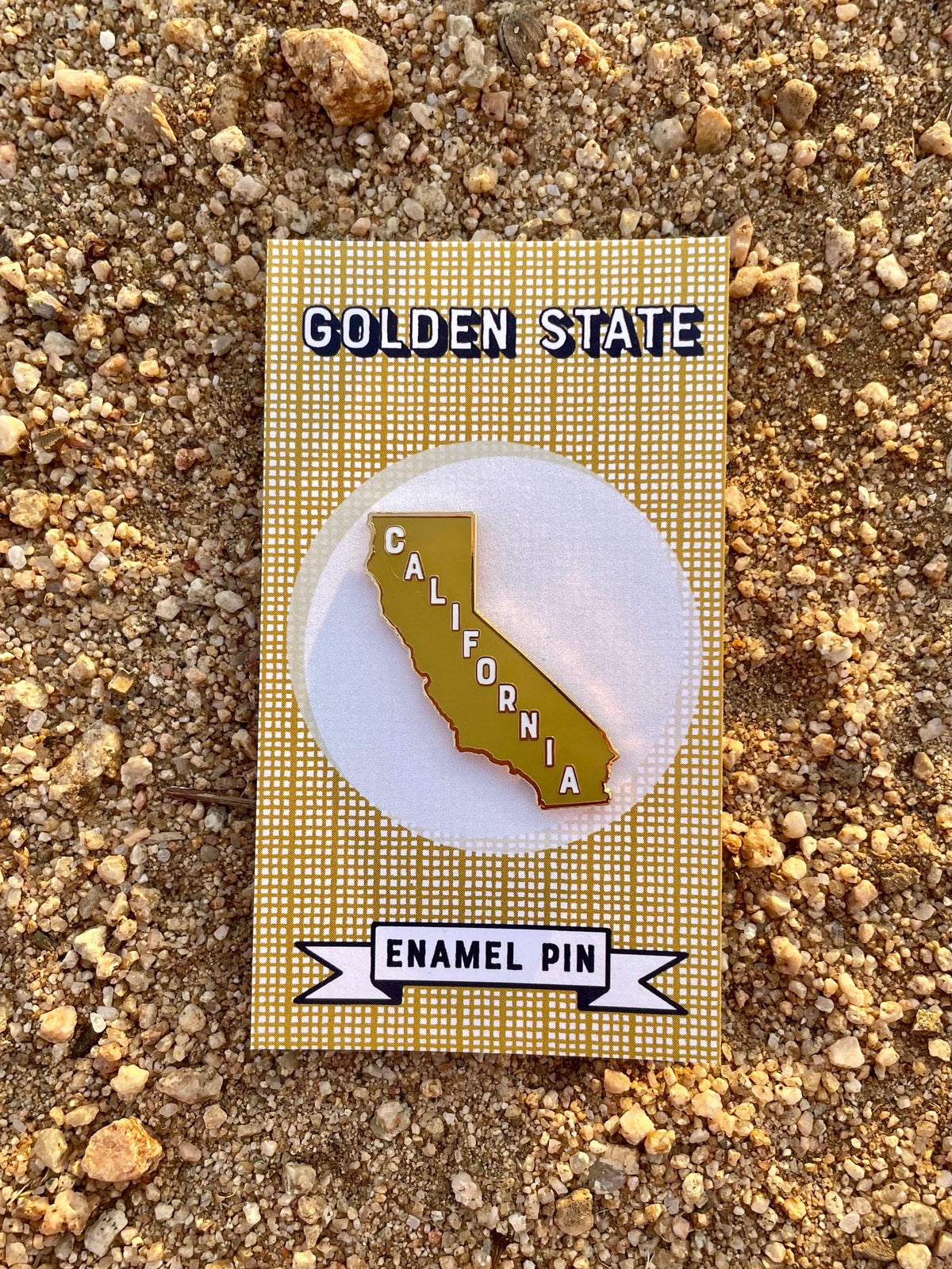 A mustard enamel pin in the shape of California on a card with the text 'Golden State' and  a dirt background.