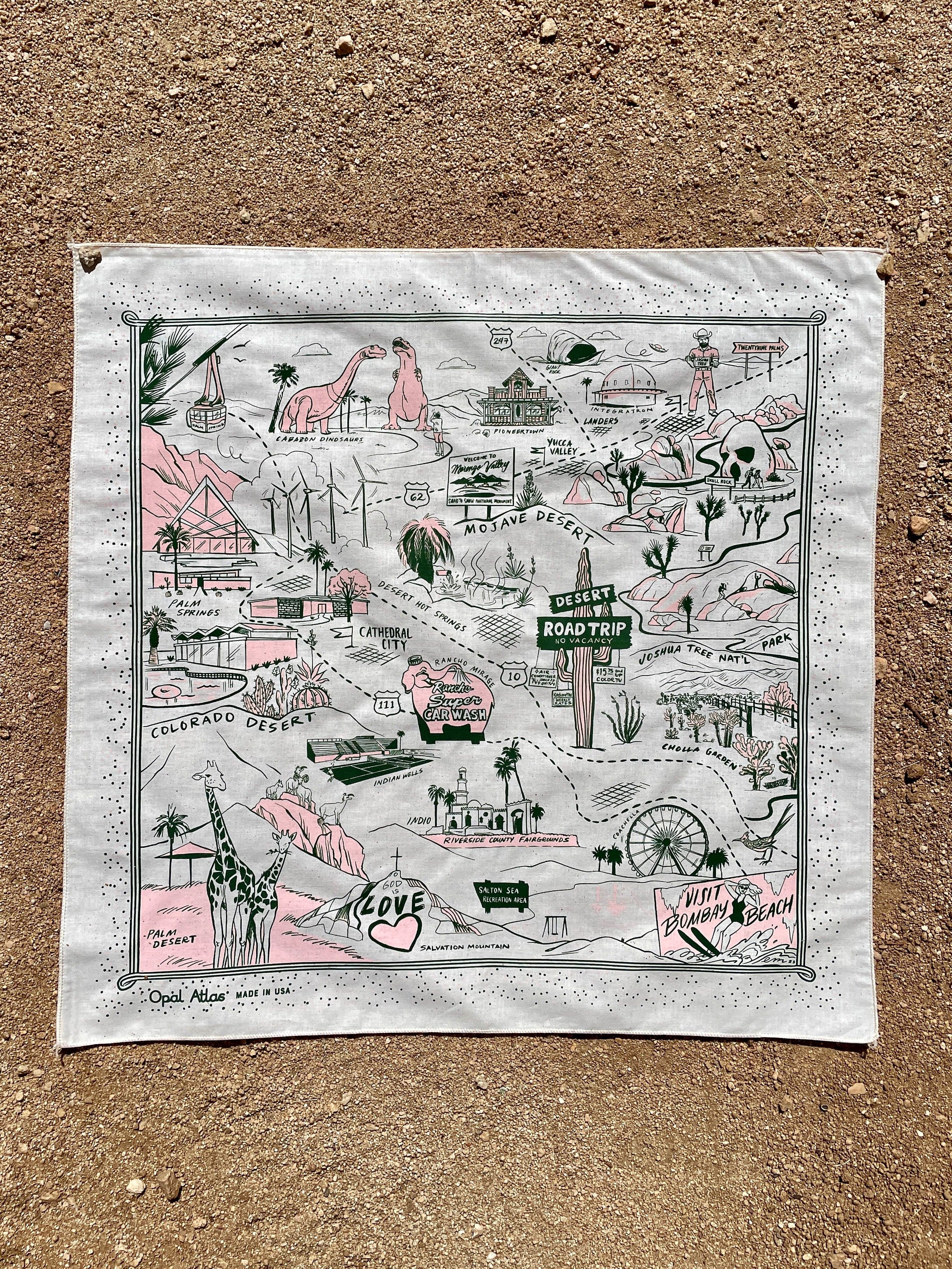 A square bandana with dark green and pink ink illustrations on a light background, laid flat on a sandy surface.