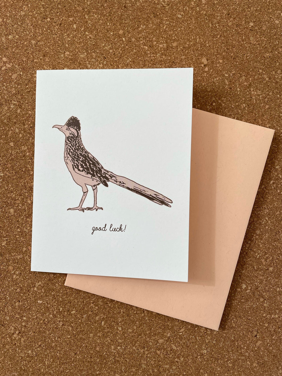 A greeting card featuring an illustration of a roadrunner with the text 'good luck!' on the front, accompanied by an orange kraft-tone envelope, all placed on a cork background.