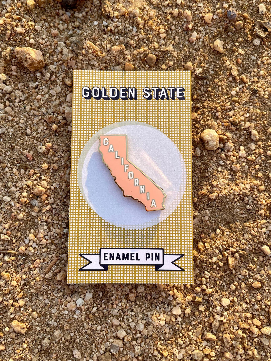 A pink enamel pin in the shape of California on a card with the text 'Golden State' and  a dirt background.