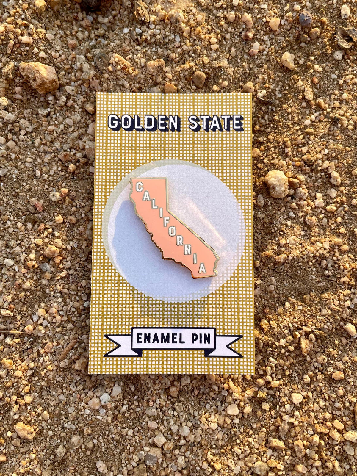 A pink enamel pin in the shape of California on a card with the text 'Golden State' and  a dirt background.