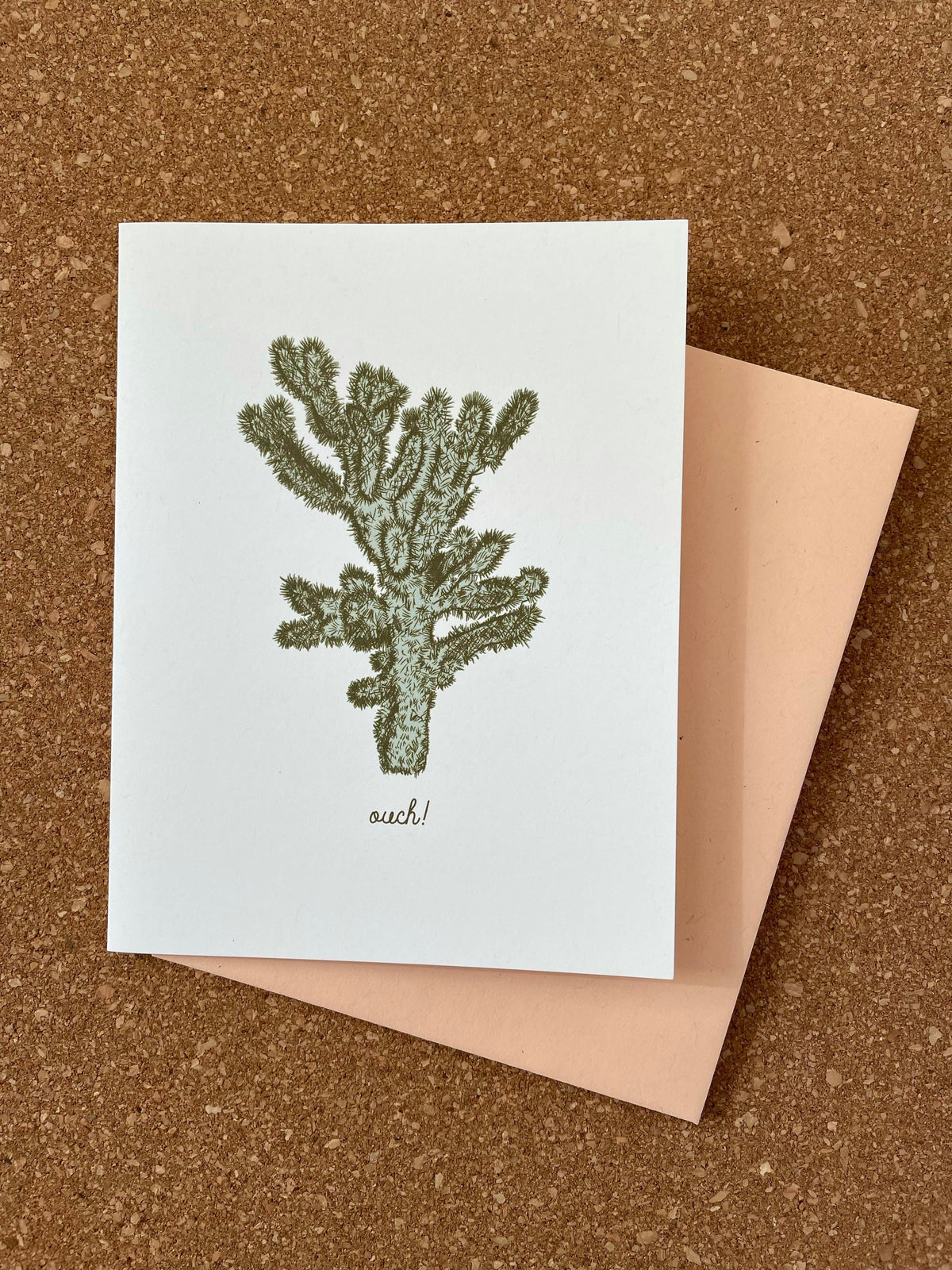 A greeting card featuring a green cactus illustration with the word 'ouch!' on it, accompanied by an orange kraft-tone envelope, all placed on a cork background.