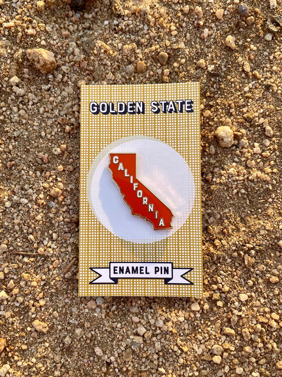 A reddish orange enamel pin in the shape of California on a card with the text 'Golden State' and  a dirt background.