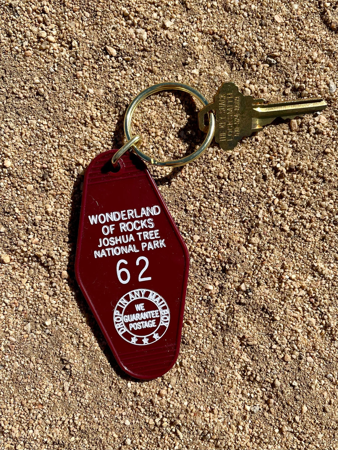 A dark red motel keychain with the text 'WONDERLAND JOSHUA TREE NATIONAL PARK 62' in white, attached to a brass keyring with a key, placed on a sandy surface.